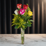 Ramo vertical de rosas rojas, rosadas, blancas y amarillas con follaje verde en un jarrón de cristal sobre mesa de mármol