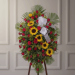 Arreglo floral tipo corona con girasoles amarillos y rosas rojas sobre base de follaje verde, colocado en caballete de madera frente a pared interior