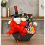 Arreglo floral AMADEA con rosas blancas, eucalipto, lavanda, vino tinto, chocolates variados y lazo rojo en cesta negra.