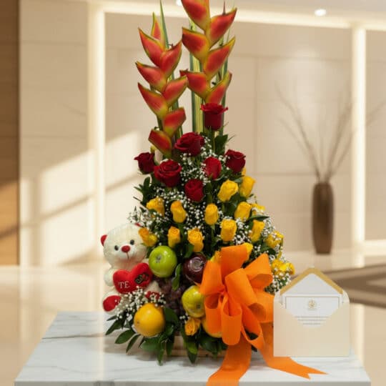 Arreglo floral con heliconias, rosas rojas y amarillas, frutas frescas, osito de peluche y lazo naranja, sobre mármol en lobb