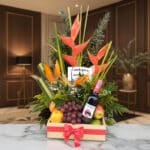 Arreglo floral Platonia con heliconias, aves del paraíso, uvas, manzanas, duraznos y botella de vino tinto, sobre mármol en u