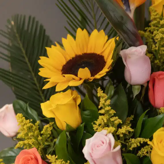 Primer plano extremo de un arreglo floral con girasol amarillo vibrante, rosas rosadas y amarillas con gotas de rocío, y foll