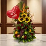 Arreglo floral premium con girasoles, rosas rojas, frutas tropicales (uvas, manzanas, naranjas) y un globo rojo de corazón co