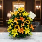 Arreglo floral tropical vibrante con girasoles, lirios amarillos y naranjas, y follaje verde, sobre una superficie de mármol