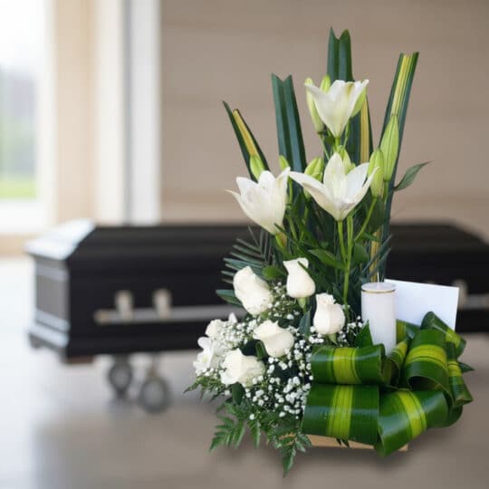 Arreglo floral fúnebre Claridad con lirios y rosas blancas, vela y tarjeta, en sala de velación moderna con ataúd al fondo.