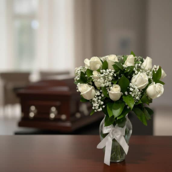 Arreglo floral fúnebre 'Pétalos del Alma' con rosas blancas frescas y follaje verde en jarrón de cristal, sobre mesa de mader