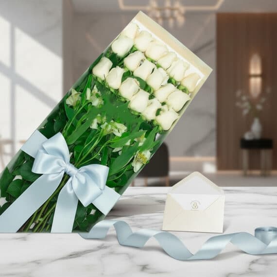 Caja de 18 rosas blancas frescas con follaje, lazo azul satinado, tarjeta de regalo y cinta decorativa sobre mármol pulido.