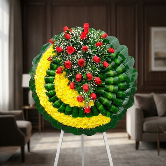Corona fúnebre Adela con rosas rojas y flores blancas sobre base de crisantemos amarillos y hojas verdes, en sala de velación