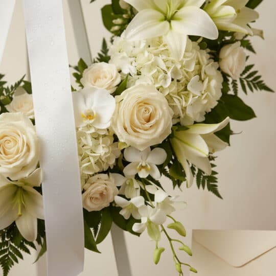 Fotografía macro premium de la Corona Fúnebre Almudena, con rosas, lirios y orquídeas blancas, follaje verde, rocío sutil y c