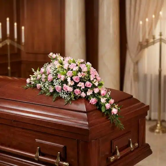 Cubre caja fúnebre elegante con rosas rosadas y follaje verde, delicadamente dispuesto sobre un ataúd de madera oscura en un