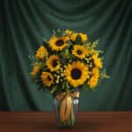 Jarrón de cristal transparente con un arreglo de 12 girasoles vibrantes, hojas verdes y pequeñas flores amarillas, atado con