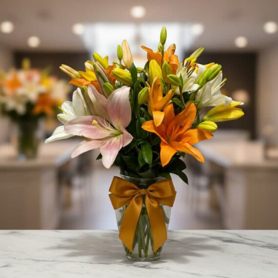 Jarrón de cristal con un moño dorado, lleno de lirios de colores variados: blancos, naranjas, amarillos y rosas, sobre una su