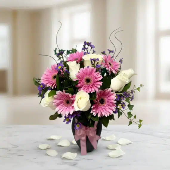 Jarrón Primavera con un elegante arreglo floral de rosas blancas, gerberas rosas y pequeñas flores moradas, sobre una mesa de