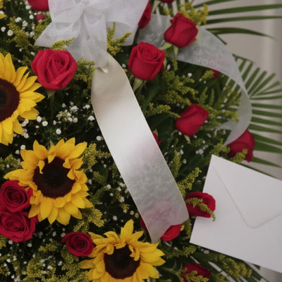 Primer plano de un pedestal fúnebre solemne con rosas rojas, girasoles amarillos y follaje verde, con una cinta blanca.